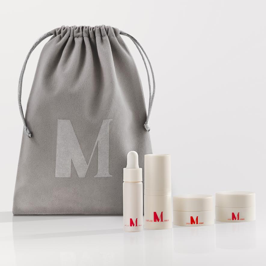Cosmetic gift set featuring an 'M' branded gray drawstring pouch with coordinated skincare bottles and containers arranged on a clean white background.